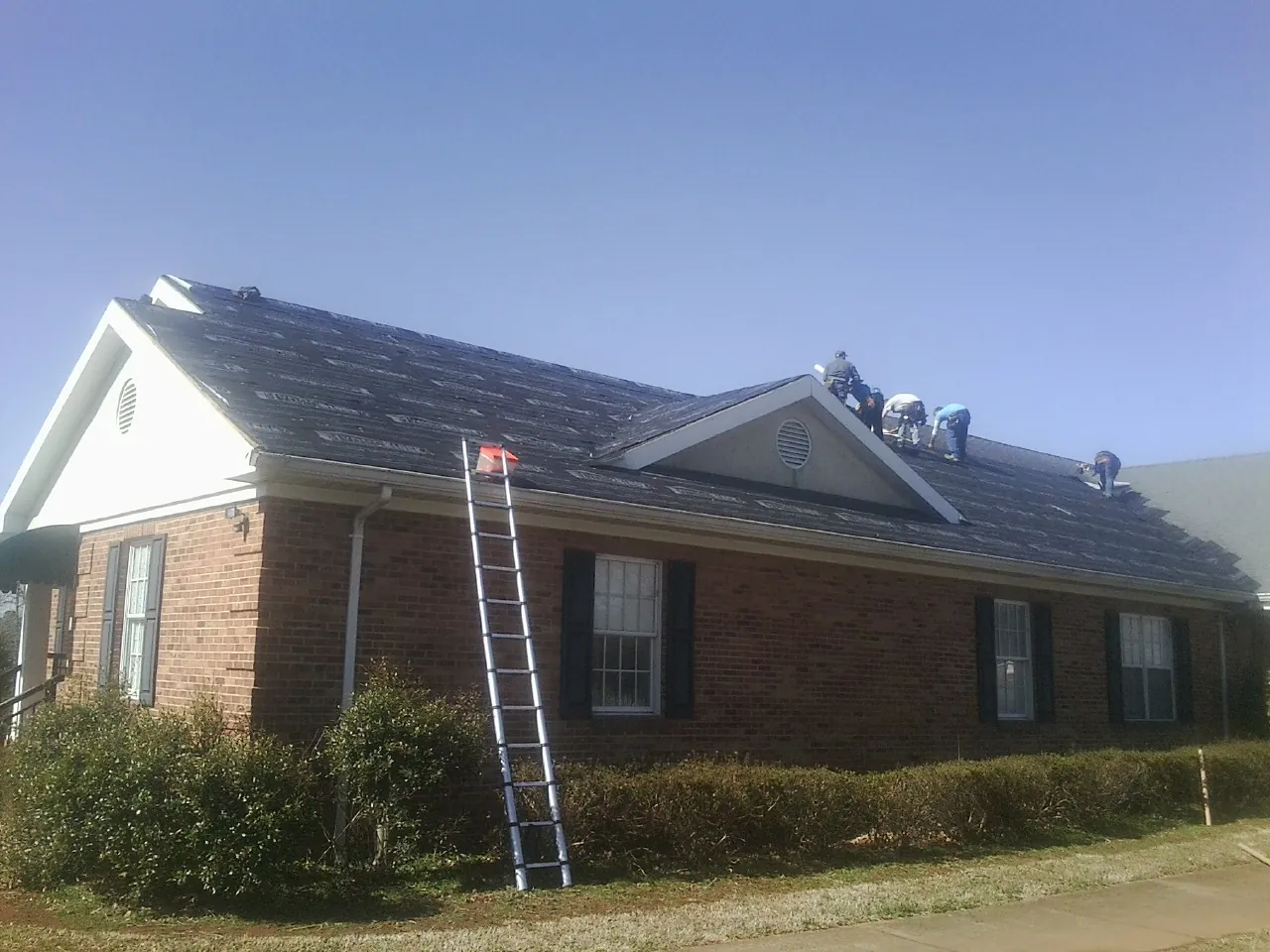 Crew tearing shingles off a brick ranch roof with a ladder set — Metal Roof Repair work by our crew in Alger