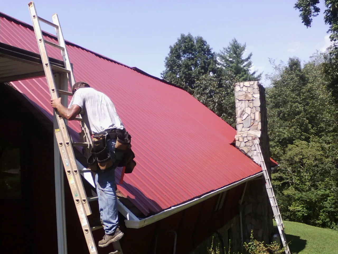 Roofer on a ladder beside a red metal roof and stone chimney — Soffit & Fascia Repair by Roofing Brothers in Alger
